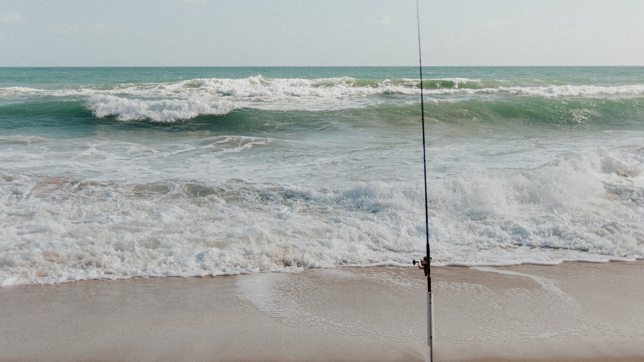 A surf fishing rod standing in a sand spike on a flat sandy Florida beach with green Atlantic surf breaking behind it
