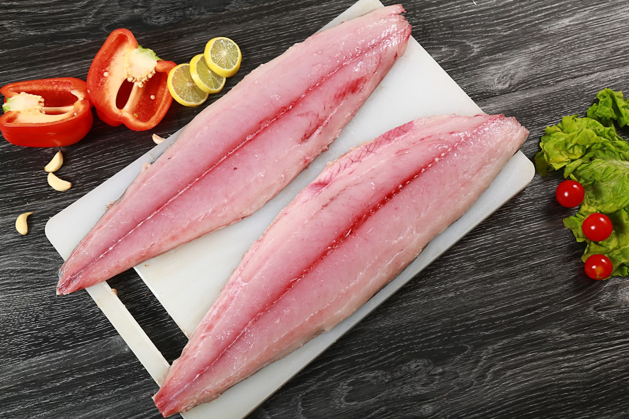 Two fresh fish fillets on a white cutting board with lemon, garlic and vegetables, bloodline visible down the centre of each fillet